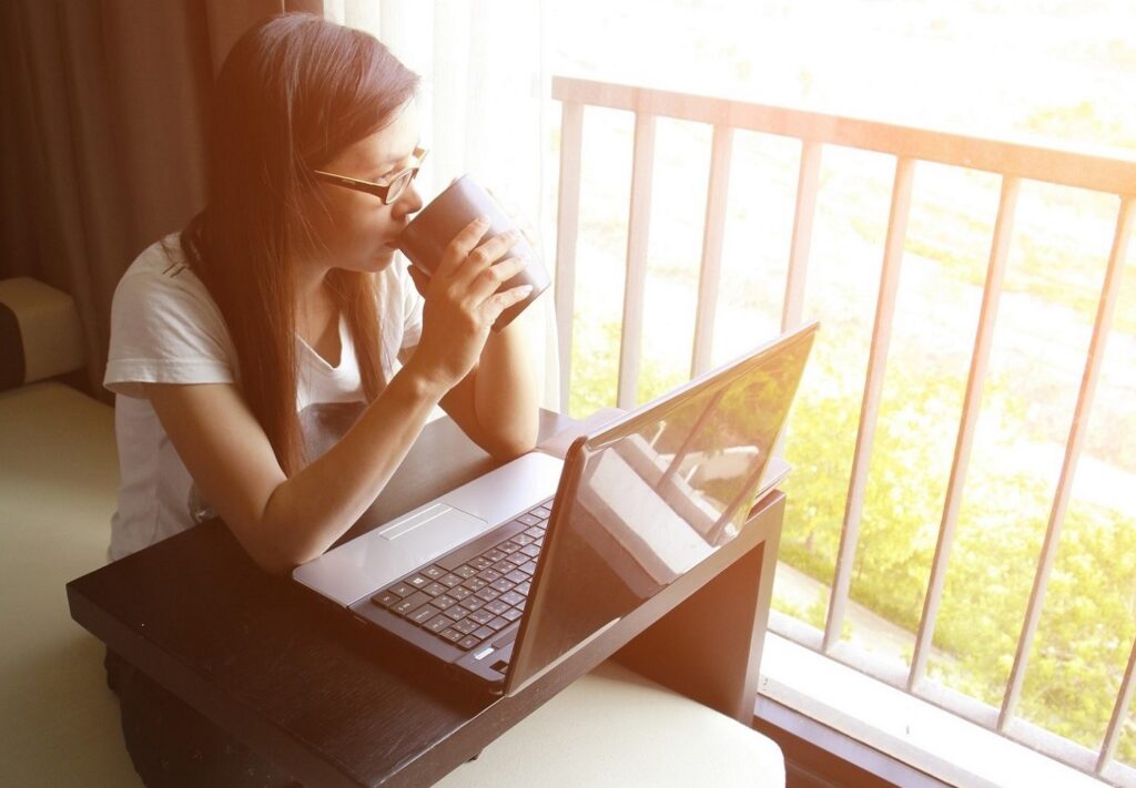 woman sitting on balcony sipping drink in front of laptop